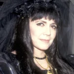 Anne Rice, the queen of gothic literature, holding a book with a mysterious background, representing her iconic vampire novels and dark romantic themes. गॉथिक साहित्य की रानी ऐन राइस की तस्वीर, जो अपनी प्रसिद्ध वैम्पायर कहानियों और अंधेरे रोमांटिक विषयों के लिए जानी जाती हैं।