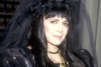 Anne Rice, the queen of gothic literature, holding a book with a mysterious background, representing her iconic vampire novels and dark romantic themes. गॉथिक साहित्य की रानी ऐन राइस की तस्वीर, जो अपनी प्रसिद्ध वैम्पायर कहानियों और अंधेरे रोमांटिक विषयों के लिए जानी जाती हैं।