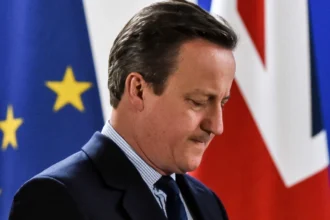 David Cameron, former UK Prime Minister, smiling in a formal suit with the UK Parliament in the background, representing his political career and life journey for Hindi and English readers on AajKyaHai.com