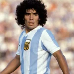 Diego Maradona in action on the football field, celebrating a goal in his iconic blue and white Argentina jersey, with fans cheering in the background.