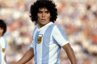 Diego Maradona in action on the football field, celebrating a goal in his iconic blue and white Argentina jersey, with fans cheering in the background.