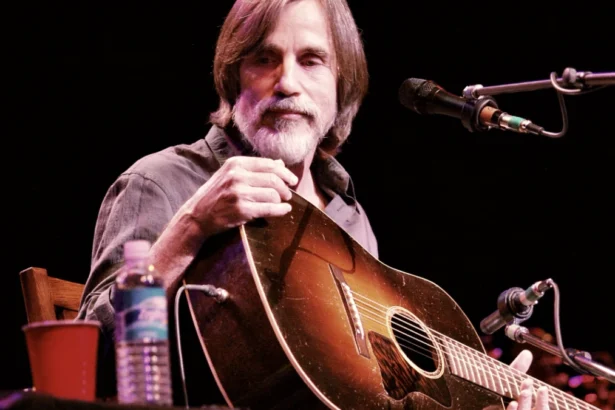 Jackson Browne smiling in a black and white portrait, holding an acoustic guitar, with musical notes and Hindi text संगीत आइकन (Music Icon) overlaid