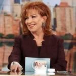 Joy Behar smiling in a studio, with Hindi text overlay 'जॉय बेहार की प्रेरणादायक जीवन कहानी' on AajKyaHai.com
