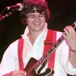 Steve Miller playing guitar on stage during a live performance, with a cheering audience in the background, showcasing his rock and blues music career.