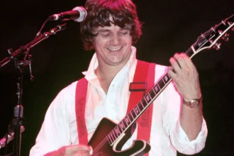 Steve Miller playing guitar on stage during a live performance, with a cheering audience in the background, showcasing his rock and blues music career.